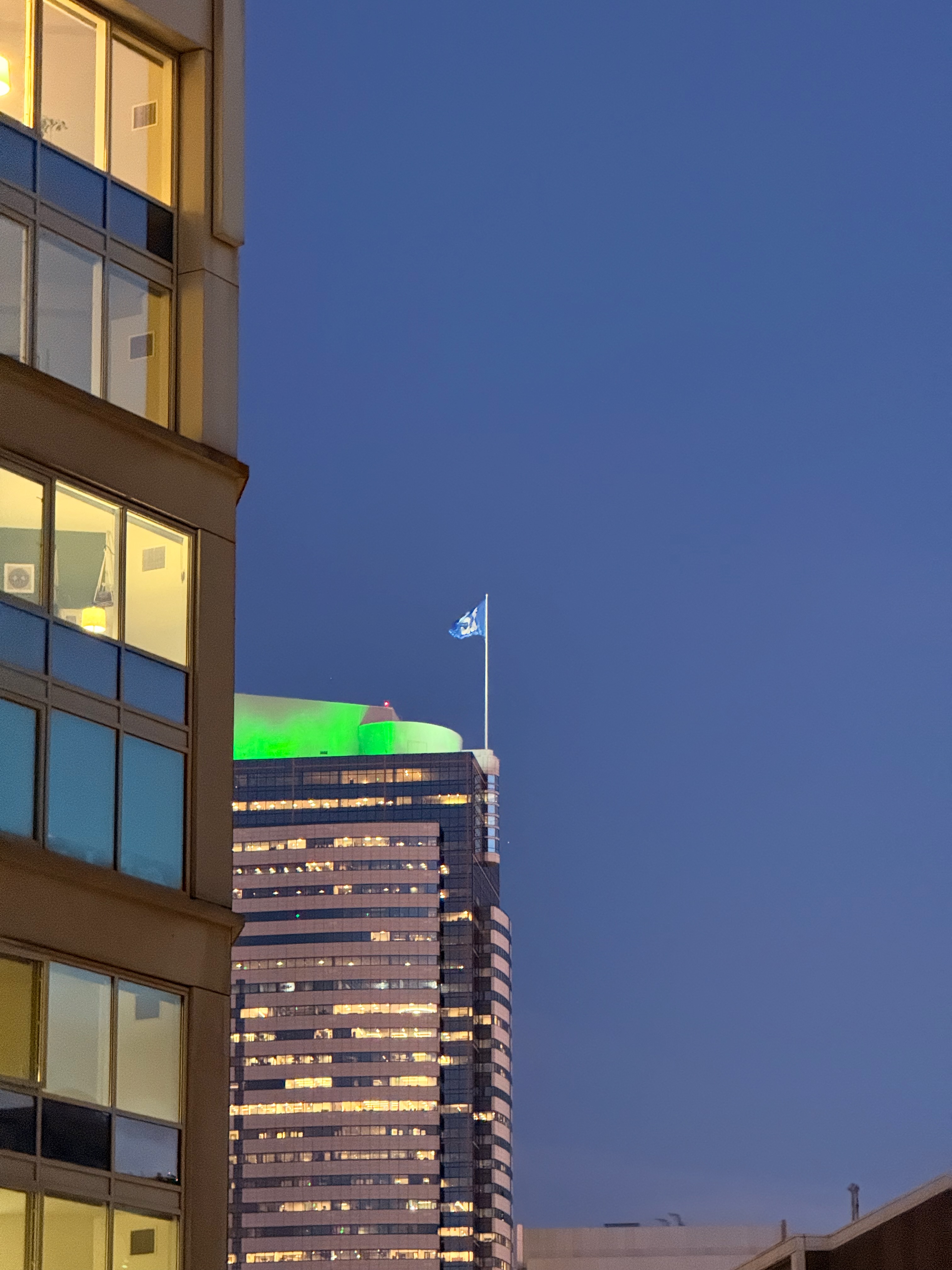 A downtown skyscraper lit up in green with a 12 flag flying on top at dusk