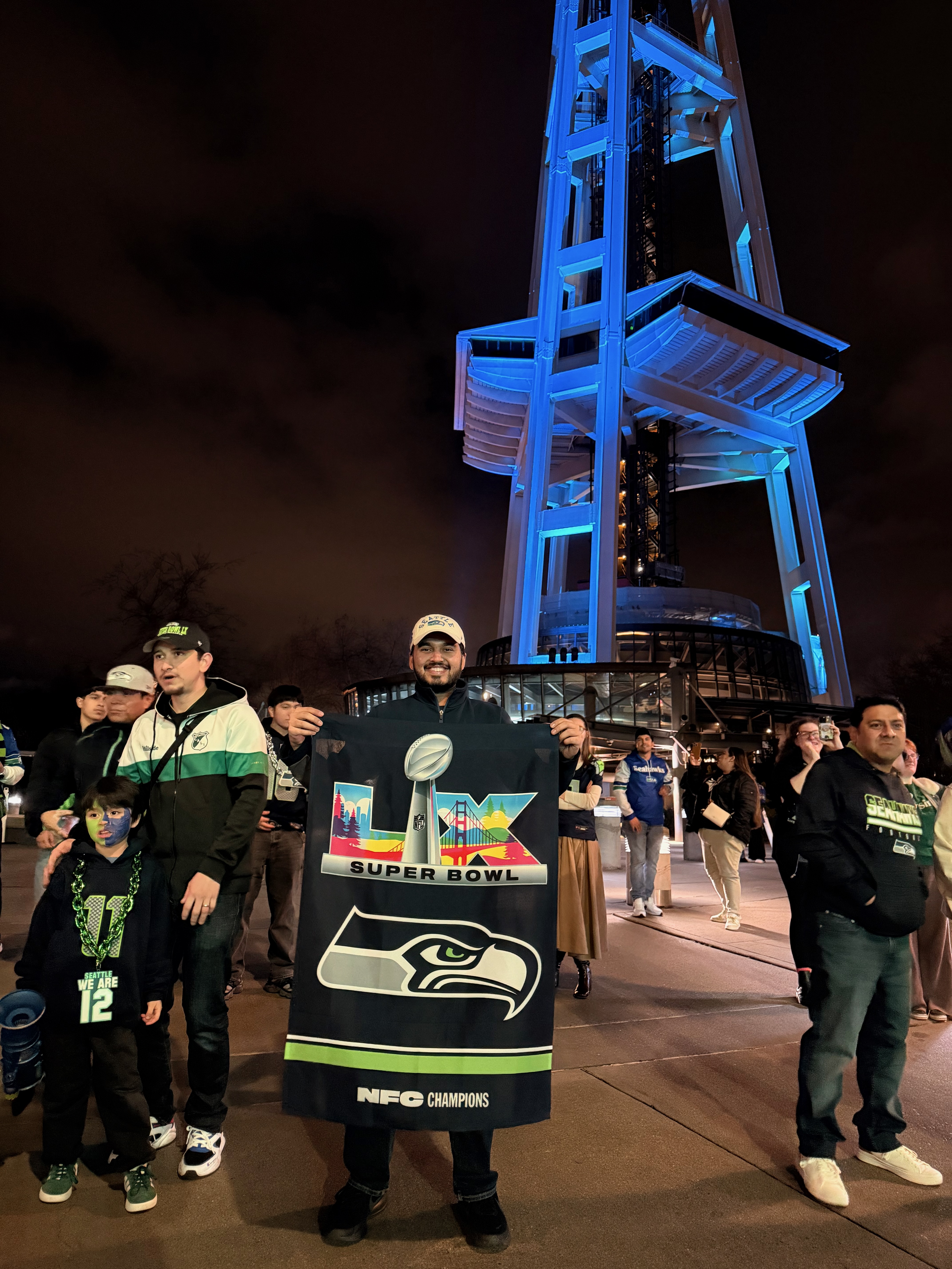 Fans celebrating at the Space Needle at night with a Super Bowl LX Seahawks banner